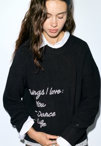 Pull noir en maille avec une coupe ample et une texture côtelée. Présente un texte manuscrit blanc : "Choses que j'aime : Toi - Danse - Vie." Superposé à une chemise blanche à col.