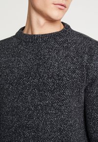 Tmavě šedý svetr s texturovaným pleteným vzorem, kulatým výstřihem a žebrovanými manžetami. Tkanina má jemný lesk a vypadá útulně.