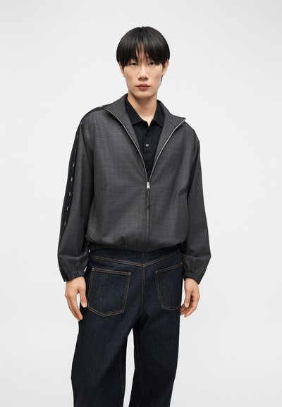 Νέος άνδρας που φοράει σκούρο γκρι ζακέτα με φερμουάρ πάνω από μαύρο πουκάμισο πόλο και σκούρα μπλε τζιν με μεγάλες πίσω τσέπες, στέκεται μπροστά σε λευκό φόντο.