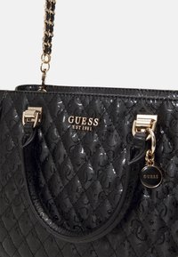 Sort quiltet håndtaske lavet af skinnende syntetisk materiale. Den har guldfarvet beslag, en kæderem og et rundt mærkeflag.