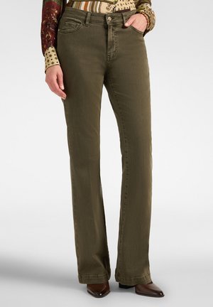 Persona che indossa pantaloni dritti verde oliva, una camicia a maniche lunghe con fantasia e scarpe marroni appuntite, in piedi con una mano in tasca.