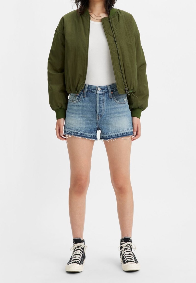 Olive green bomber jacket with ribbed cuffs, white fitted tank top, and distressed denim shorts. Black high-top sneakers complete the look.