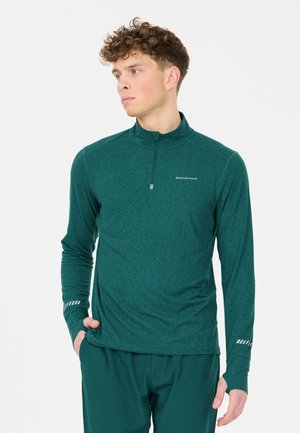 Jonge man met krullend haar draagt een teal-kleurige sporttop met lange mouwen en een kwart rits, kijkt zijwaarts met een hand in zijn zak.