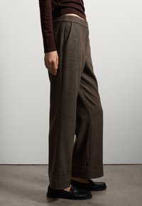 Pantalons en laine marron à jambes larges avec une finition texturée, dotés de plis avant et d'ourlets retournés. Associés à des mocassins noirs.