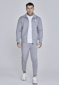 Giacca zip grigia chiara e pantaloni affusolati abbinati, realizzati in tessuto morbido, con logo sul petto e sull'anca, abbinati a sneakers bianche.