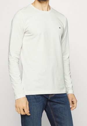 Mann trägt ein schlichtes weißes Langarmshirt und blaue Jeans, steht vor hellem Hintergrund, mit kleinem Logo auf der Brust.