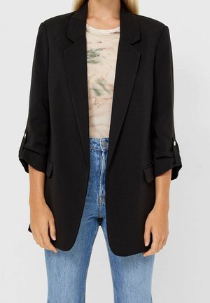 Femme portant un blazer noir aux manches retroussées, une chemise beige à motifs et un jean bleu taille haute sur un fond uni.