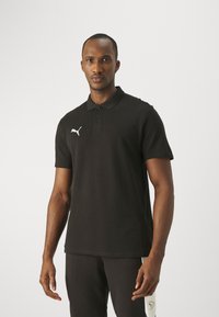 Čierna polo košeľa z mäkkej tkaniny s klasickým golierom, tromi gombíkmi a bielym logom Puma na ľavej strane. Nosená s čiernymi nohavicami.