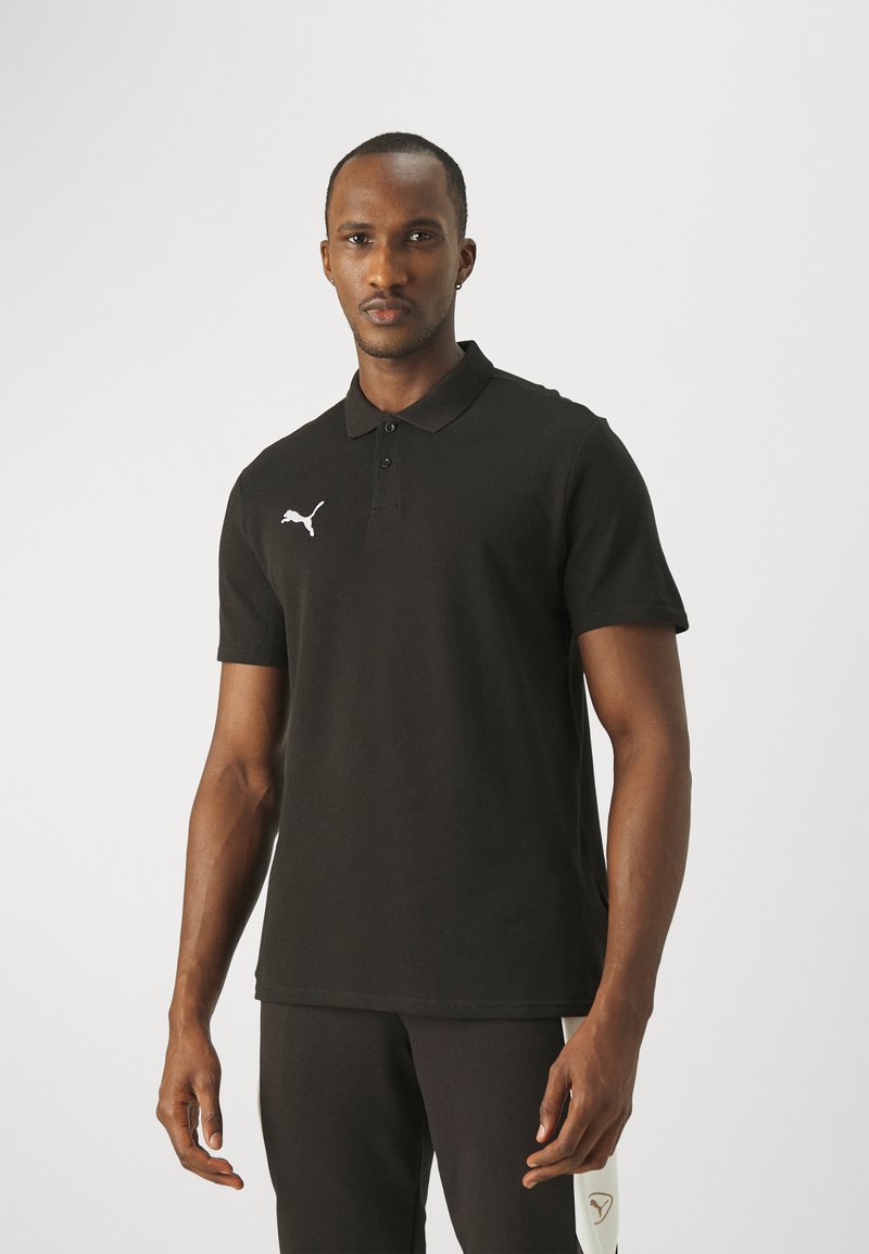 Čierna polo košeľa z mäkkej tkaniny s klasickým golierom, tromi gombíkmi a bielym logom Puma na ľavej strane. Nosená s čiernymi nohavicami.