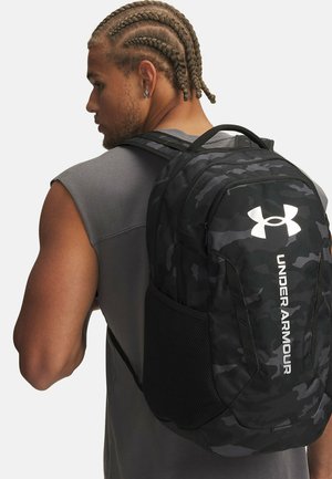 Mustast kamuflaažimustriga seljakott võrgust küljetaskute, reguleeritavate rihmade ja valge "UNDER ARMOUR" logoga ees. Vastupidav kangasmaterjal.
