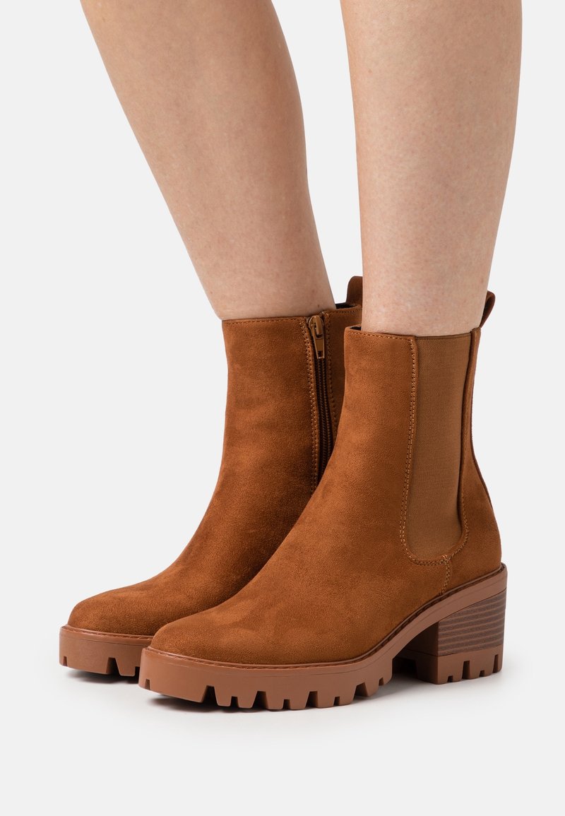 Bottines en daim marron avec talon bloc, dotées de panneaux latéraux élastiques et d'une fermeture éclair arrière. Semelle en caoutchouc texturée avec rainures de traction.