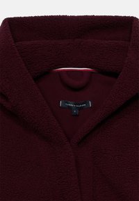 Μπουφάν από μπορντό φλις με γιακά σαν ριχτόκαμπο, χαρακτηριστικό απαλής υφής. Η εσωτερική ετικέτα δείχνει την επωνυμία «Tommy Hilfiger» και το μέγεθος «S».