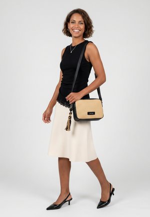 Lachende vrouw met een zwarte mouwloze top, een crèmekleurige rok, zwarte slingback hakken, met een beige en zwarte crossbodytas met tassel-detail.