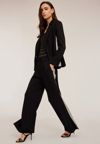 Americana negra ajustada con cierre de un solo botón, combinada con pantalones negros sueltos con rayas blancas a los lados y una blusa de rayas negras.