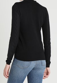 Schwarzer Strickpullover mit langen Ärmeln im Spitzenmuster. Rippkragen und -saum. Kombiniert mit hellblauen Jeans.