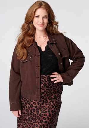 Vrouw met lang rood haar die een bruine jas met studs draagt, een zwart kanten top, een rok met luipaardprint en gelaagde gouden kettingen, glimlachend met haar hand op haar heup.
