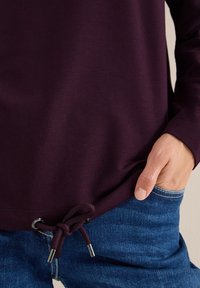 Bordeaux sweatshirt med lange ærmer og snøre i kanten. Glat tekstur med sølvfarvede hardwaredetaljer. Sammen med blå jeans.