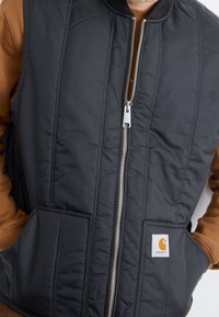 Černá prošívaná vesta s vysokým límcem, předním zipem a dvěma bočními kapsami. Na dolní kapse se nachází logo Carhartt.