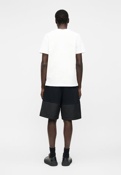 Άνδρας που στέκεται με την πλάτη, φοράει μονόχρωμο λευκό κοντομάνικο μπλουζάκι, μαύρο φαρδύ σορτς, μαύρες κάλτσες και μαύρα αθλητικά παπούτσια σε λευκό φόντο.