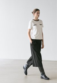 Femme marchant à l'intérieur portant un haut à manches courtes blanc avec un liseré noir, un pantalon large noir raccourci et des bottes noires montant jusqu'aux genoux.