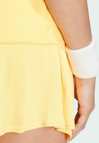 Vestito sportivo giallo con tessuto testurizzato, gonna plissettata e etichetta ricamata "HEAT RDY". Accompagnato da un polsino in terry bianco.