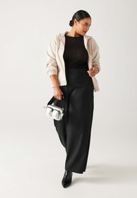 Femme portant un hoodie beige zippé, un haut texturé noir, un pantalon large noir, des bottes noires, et tenant un petit sac à main argenté métallique.