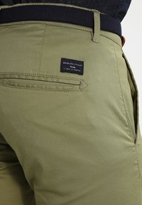 Olive green pants with a textured finish, featuring a black waistband and a back pocket with a small brand label.