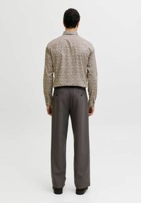 Homme de dos portant une chemise à manches longues à motif floral rentrée dans un pantalon large gris et des chaussures noires sur un fond uni.