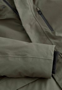 Giacca da esterno verde scuro in tessuto con cucitura orizzontale, dettaglio ai polsini elasticizzati e tasca con zip nera. Texture liscia e design funzionale.