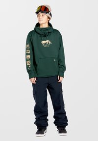Zielona bluza z kapturem z złotą grafiką i napisem, regulowanym sznurkiem i przednią kieszenią. Połączona z granatowymi spodniami cargo i czarnymi sneakersami.