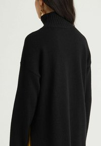 Pull en maille noire avec un col montant côtelé et des coutures d'épaule tombantes. Il présente une texture douce et une coupe décontractée.