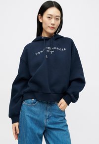 Γυναίκα που φοράει μια ναυτική μπλε κουκούλα Tommy Hilfiger και μπλε τζιν, στέκεται με το ένα χέρι στην τσέπη μπροστά σε έναν απλό λευκό φόντο.