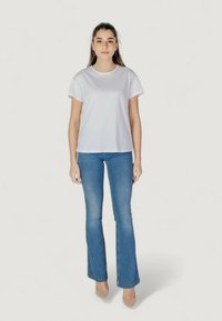 Camiseta de algodón blanca de manga corta, combinada con unos vaqueros de campana azules. El modelo está de pie contra un fondo liso.
