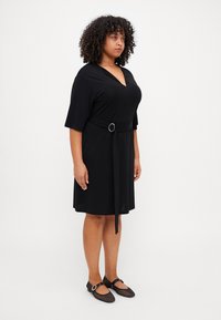 Černé šaty ke kolenům s výstřihem do V a krátkými rukávy, které mají látkový pásek s kulatou sponou. Spárovány s černými síťovanými balerínkami.