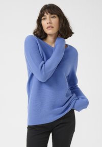 Blauer gerippter Strickpullover mit lockerer Passform und weitem Halsausschnitt. Der Stoff hat eine glatte Textur und lange Ärmel, kombiniert mit schwarzen Hosen.