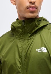Close-up af en person iført en olivengrøn lynlåstrøje med The North Face-logo på brystet, der viser kraven og en del af ansigtet.