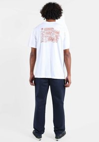 T-shirt blanc à manches courtes avec un motif corail graphique dans le dos, associé à un pantalon bleu marine et des chaussures de skate noires.