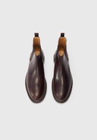 Une paire de bottes Chelsea en cuir brun foncé poli avec des languettes et des panneaux élastiques sur un fond blanc.