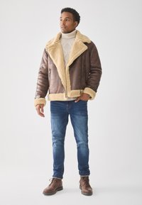 Chaqueta de cuero marrón con forro de borreguillo beige, cierre de cremallera en la parte delantera y cuello ancho; combinada con un suéter de punto trenzado color crema y jeans azules.