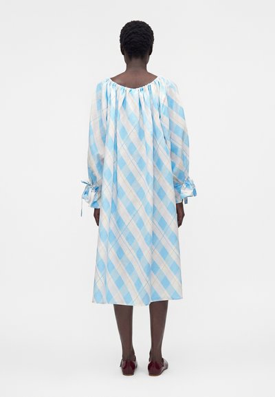 Άτομο που φορά χαλαρό, μακρυμάνικο μπλε και λευκό καρό φόρεμα με σούρες στο ντεκολτέ και δεμένα μανίκια, στέκεται με την πλάτη.
