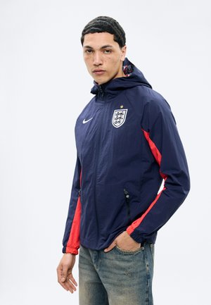 Młody mężczyzna ubrany w granatową kurtkę piłkarską Anglii z czerwonymi paskami i dżinsy, pozujący z jedną ręką w kieszeni na białym tle.