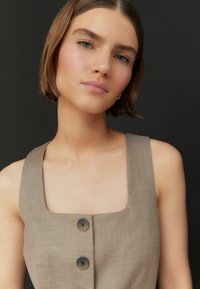 Abito beige con scollo quadrato e due bottoni scuri sul davanti, realizzato in un tessuto testurizzato. Il modello ha capelli di lunghezza medio-lunga e trucco neutro.