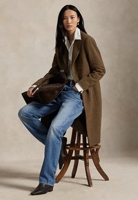 Woman wearing brown coat, white shirt, blue jeans, and black boots, sitting on wooden stool holding dark brown bag against neutral background.