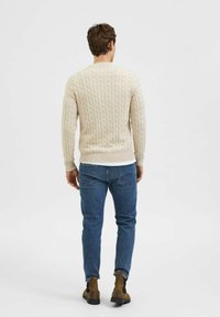 Suéter beige de punto trenzado con puños y dobladillo acanalados, combinado con jeans de mezclilla azules y botas marrones hasta el tobillo, visto desde atrás.