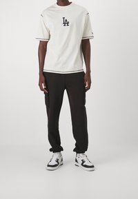 Krémové tričko s černým logem "LA", černým lemem, v kombinaci s černými cargo kalhotami a černo-bílými teniskami s šněrováním.