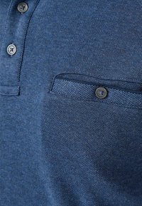 Lyhytsleeve polo-paita tummansinisestä kankaasta, jossa on teksturoitu kudos. Takana rintatasku ja pyöreät napit, jotka tarjoavat rentoja suunnitteluelementtejä.