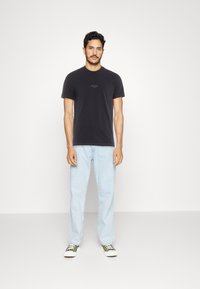 Black cotton t-shirt with a small white "GUESS" logo on the chest, paired with light blue denim jeans and green sneakers.
