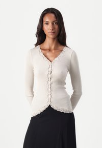 Beige ribbed knit cardigan with long sleeves, featuring a scalloped lace trim and button closure down the front. Smooth texture, fitted design.