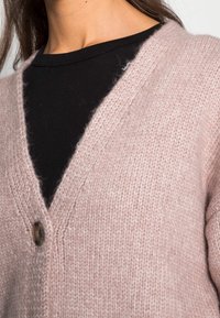 Světle růžový pletený cardigan s výstřihem do V a jedním kulatým knoflíkem, s měkkou texturovanou látkou přes černé tričko.