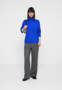 Blue cable-knit turtleneck sweater paired with gray pinstriped wide-leg trousers. Model holds a metallic handbag; black shoes complete the look.